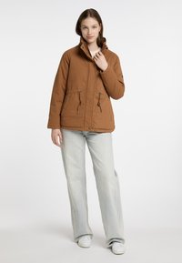 Brun isoleret jakke med høj krave, snor i taljen og knappet front. Kombination med lyseblå flared jeans og hvide sneakers.