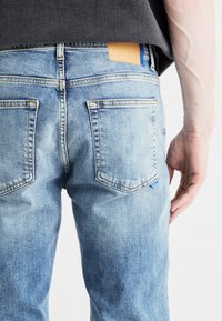 Jeans de mezclilla azul claro con un acabado desgastado, que cuentan con una etiqueta de cuero marrón en la cintura y dos bolsillos traseros.