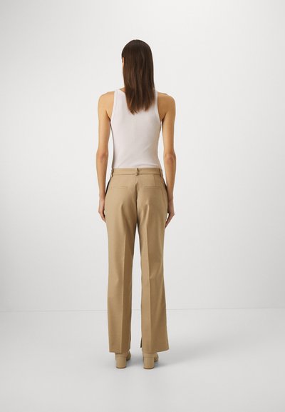 Beige skreddersydde bukser med en glatt tekstur, høyt liv og to baklommer. Brukt med en tettsittende hvit tanktop og beige sko.
