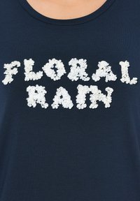 Námořnická modrá tričko s textem "FLORAL RAIN" složeným z bílých aplikací s květinovým vzorem v texturovaném, prostorovém designu.