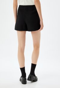Shorts noirs texturés avec une ceinture lisse, longueur mi-cuisse, dotés de coutures distinctives. Portés avec des chaussettes et des chaussures noires.