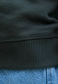 Fekete pamut bordás pulóver alja texturált felülettel, világoskék farmerrel párosítva, amelyen varrási részletek láthatók.