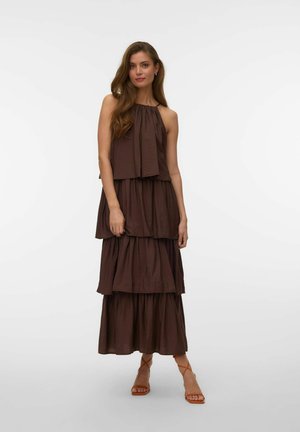 Femme aux cheveux longs portant une robe maxi marron foncé sans manches et à volants, ainsi que des sandales à lanières couleur fauve, debout devant un fond uni.