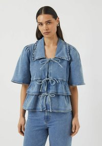 Paita denim peplum dengan lengan mengembung, warna biru muda, bagian depan kancing, pinggang dengan tali serut, dan tepi kerah yang bergerigi. Kain bermutu tekstur.