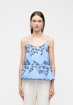 Γυναίκα που φοράει μπλε αμάνικη μπλούζα με σκούρα κεντήματα λουλουδιών και τελείωμα με κυματιστό σχέδιο, συνδυασμένη με ανοιχτό μπεζ παντελόνι και κρεμαστά σκουλαρίκια.