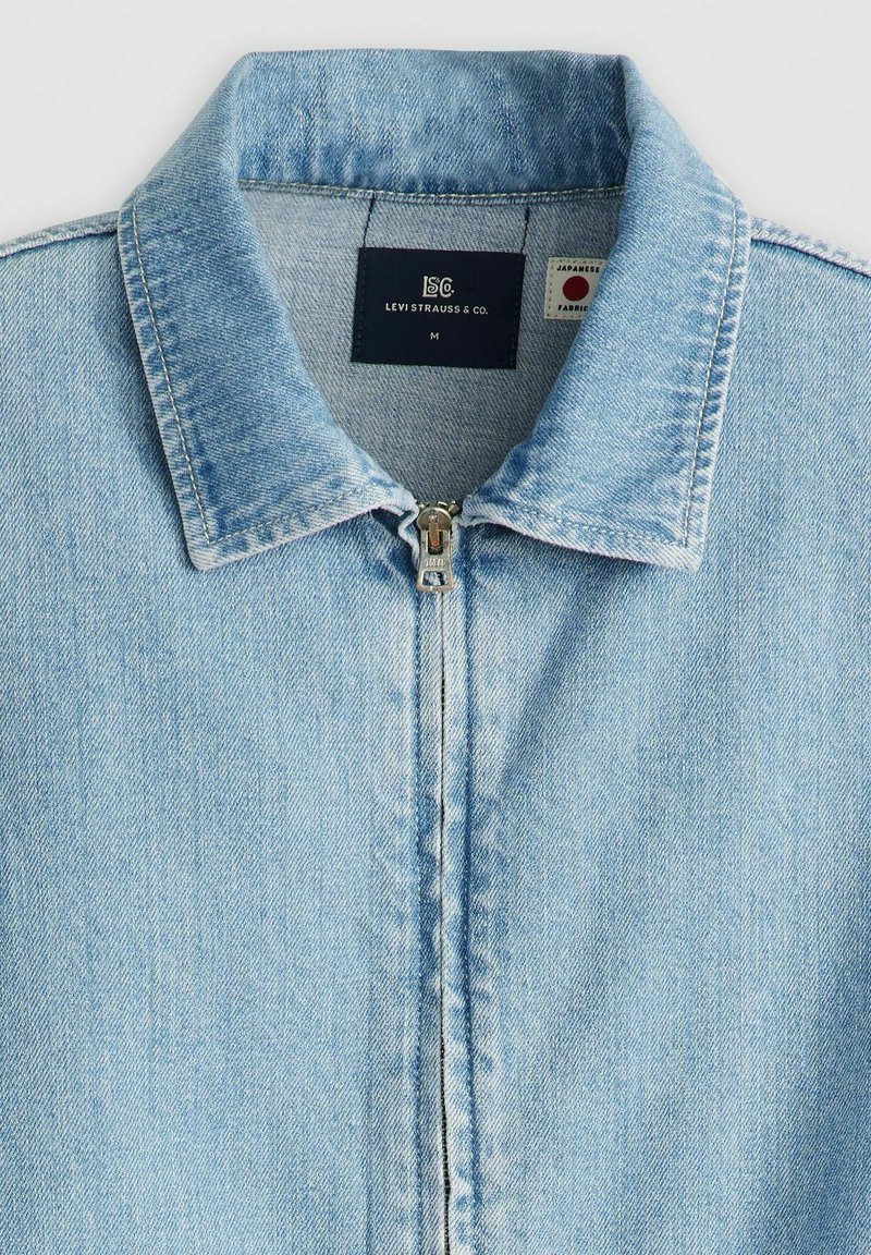 Hellblaues Jeansjacke mit einem Reißverschluss, spitzem Kragen und vertikalen Nahtdetails. Mit Markenetikett im Nacken.