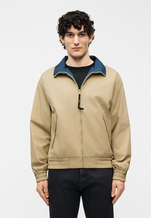 Ung mand med krøllet hår iført en beige jakke med lynlås og blå krave samt sorte bukser, stående foran en hvid baggrund.