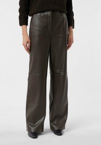 Wide-leg trousers in dark brown leather. Smooth texture with a high waist and creased detailing. Paired with black heeled shoes.