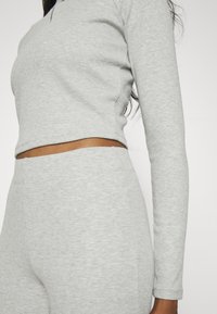 Donna che indossa un top corto a maniche lunghe a coste grigio chiaro e pantaloni a coste abbinati a vita alta, mostrando il ventre e il braccio.