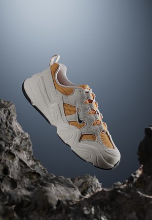 Chaussure de sport blanche et jaune moutarde épaisse avec textures en mesh et suède, semelle extérieure noire et petit logo Swoosh noir, flottant au-dessus d'une surface rocheuse.