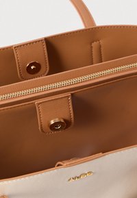 Intérieur d'un sac à main en cuir tan présentant une poche zippée, un fermoir à pression et des accents en métal doré. L'extérieur arbore une couleur claire contrastante.