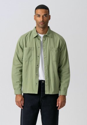 Muž oblečený v svetlozelené košeli na gombíky cez bielu košeľu a čierne nohavice, stojaci pred jednoduchým svetlosivým pozadím.