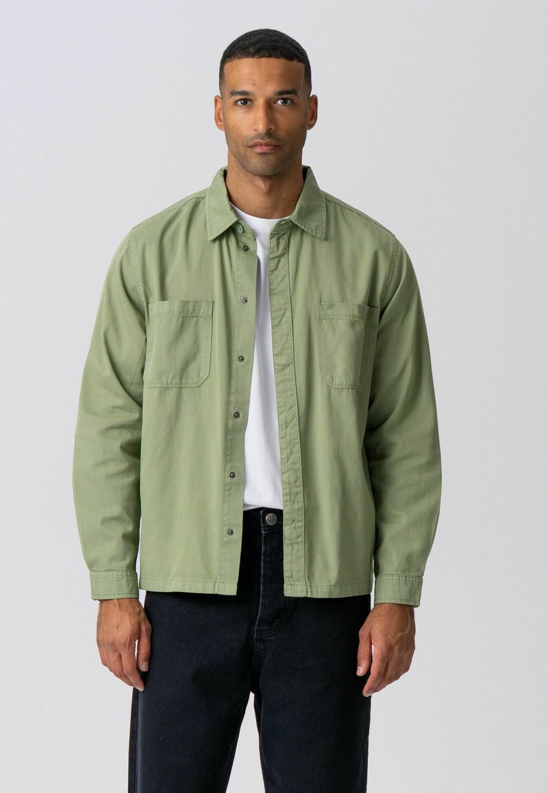Homme portant une surchemise boutonnée vert clair par-dessus une chemise blanche et un pantalon noir, debout devant un fond gris clair uni.