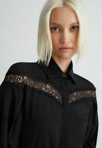 Camicia nera con colletto, caratterizzata da pannelli in pizzo, bordi sfrangiati sulle spalle e chiusura con bottone al colletto. Tessuto dalla texture liscia.