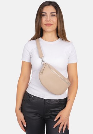 Beige Leder-Crossbody-Tasche mit geschwungenem Design, ausgestattet mit einem silbernen Reißverschluss und einem Quastenakzent, getragen über einem weißen Hemd und schwarzen Hosen.