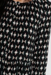 Černé šaty se vzorem s geometrickými tvary v růžové, tyrkysové a krémové barvě, s knoflíkovým zapínáním a staženým detailem v pase.