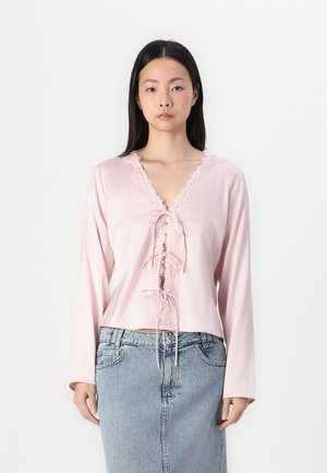 Pink satin blouse featuring a V-neckline with lace trim and ribbon detailing at the center. Long sleeves. Worn with a denim skirt.