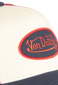 Krēmas un tumši zils cepure ar sarkanu izšūtu "Von Dutch" logotipu uz ovālas plāksnes. Iekļauj šuvju detaļas un strukturētu pārklājumu.