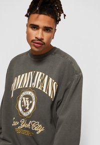 Felpa grigia realizzata in tessuto morbido con la scritta "TOMMY JEANS" in grassetto giallo e un logo tondo con la scritta "New York City".