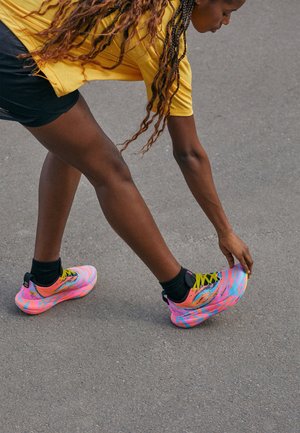 Woman in yellow shirt and black shorts stretching leg, touching colorful pink and blue running shoe on gray pavement.