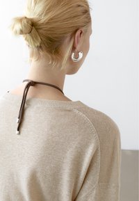 Maglione beige a maglia con una vestibilità rilassata, dotato di scollatura tonda e dettaglio con fiocco marrone sul retro. Areeggetti grandi in argento a cerchio visibili.