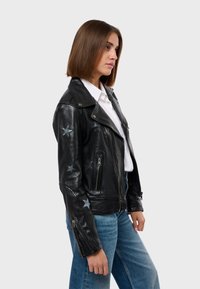 Crna kožna motoristička jakna sa srebrnim zvjezdastim detaljima, dzepovima na patentni zatvarač i rebrastim ovratnikom, nosi se preko bijele košulje i plavih traperica.