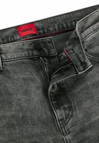 Pantaloni in denim grigio con etichetta rossa con la scritta "HUGO", chiusura con zip e bottone. Il tessuto ha una finitura testurizzata e un motivo sottile.