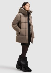 Woman wearing a taupe padded coat with high collar, black dress, black tights, and black lace-up winter boots, standing against a plain background.