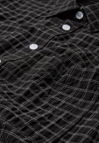 Tissu noir à motif quadrillé blanc avec des boutons ronds blancs alignés verticalement sur une chemise froissée.