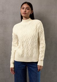 Jeune femme aux longs cheveux noirs, portant un pull en maille texturé crème et un jean bleu, se tenant contre un mur en béton gris.