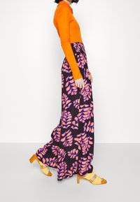 Kvinde der går iført en orange langærmet top, brede sorte bukser med pink bladmønstre og gule højhælede sandaler mod en hvid baggrund.