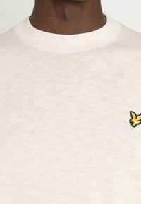 Pull beige clair à col rond en tissu côtelé doux. Présente un petit logo jaune brodé sur la partie gauche de la poitrine.