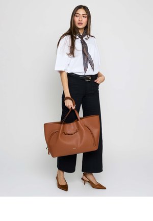 Mujer con cabello largo y oscuro que lleva blusa blanca, pantalones negros, pañuelo estampado en el cuello, tacones marrones y sostiene un gran bolso tote de cuero marrón.