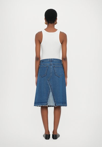 Fustă din denim cu margini desfăcute, până la genunchi, cu un design cu fendă și două buzunare pe spate, purtată cu un top alb fără mâneci și balerini negri.