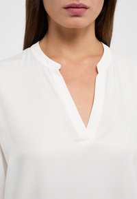 Weiße Bluse mit V-Ausschnitt, aus weichem Stoff, glatter Textur und minimalistischem Design, das klare Linien und Einfachheit betont.