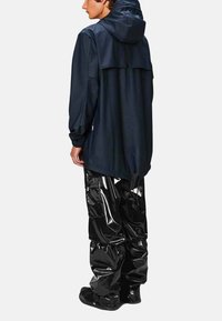 Veste imperméable navy avec capuche et poignets élastiques, assortie à un pantalon en vinyle noir brillant. Les deux pièces présentent des designs amples et oversize.