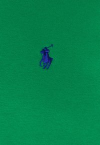 Grönt bomullstyg med en liten, broderad lila polo-spelare-logo på vänster sida. Texturen är slät och lätt.