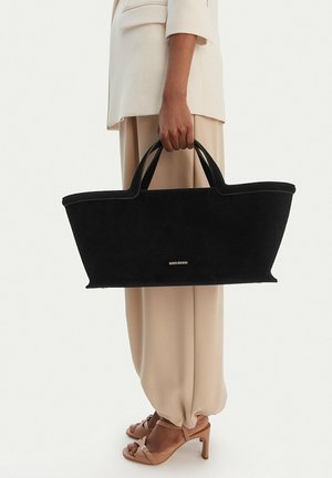 Osoba v béžovém obleku a na podpatcích držící velkou černou tašku přes rameno s logem "GINO ROSSI" za úchyty po straně.