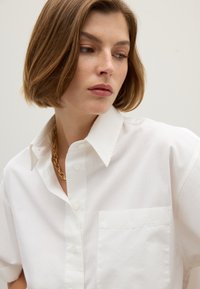 White cotton shirt with a classic collar, short sleeves, a chest pocket, and a relaxed fit, featuring button closure and slight sheen.