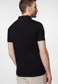 Czarna koszulka polo wykonana z teksturowanej tkaniny, z krótkimi rękawami i kołnierzykiem. Zauważalny czerwony detal logo na karku.