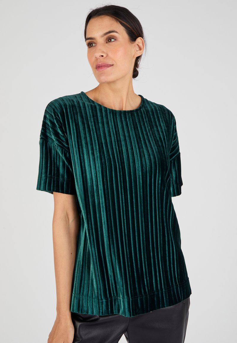 Top de terciopelo verde con rayas verticales, mangas cortas y cuello redondo. Textura suave con un corte holgado, combinado con pantalones oscuros.