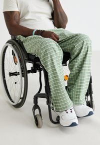 Groene geruite broek van lichtgewicht stof, gedragen door een persoon in een rolstoel. Witte sportschoenen met blauwe zolen en kleurrijke accenten.