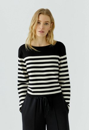 Vrouw met blond haar die een zwart-wit gestreept top met lange mouwen en een zwarte broek draagt tegen een lichtgrijze achtergrond.