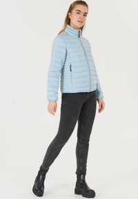 Lysblå puffjakke med høj krave, vandret syninger, to lynlåslommer i siderne, parret med sorte tætsiddende jeans og ankelsko.