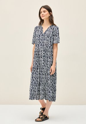 Vestido maxi de algodón navy con patrones geométricos blancos, mangas cortas, escote en V y falda de volantes. Combinado con sandalias negras con hebillas doradas.