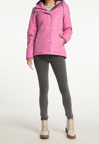 Kobieta w różowej kurtce z kapturem, ciemnoszarych dopasowanych dżinsach i beżowych sznurowanych botkach na kostkę stoi na neutralnym tle.