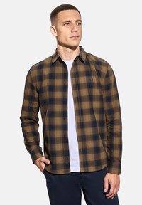 Muž v hnědém a černém kostkovaném košilovém triku přes bílém tričku s námořnickými kalhotami, stojící s jednou rukou v kapse.