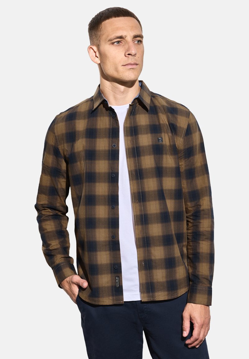 Muž v hnědém a černém kostkovaném košilovém triku přes bílém tričku s námořnickými kalhotami, stojící s jednou rukou v kapse.