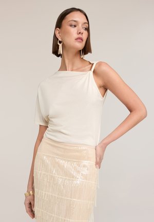 Lyhyttukkainen ruskeahiuksinen nainen, jolla on yläosassa yksipuolinen kerma paita ja beige paljettihapsuhame, asusteena kultaiset korvakorut ja rannekorut.