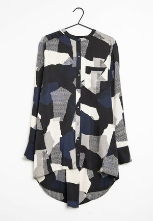 Chemise à manches longues boutonnée avec un motif patchwork abstrait irrégulier en noir, blanc, gris et bleu marine, suspendue sur un cintre noir.